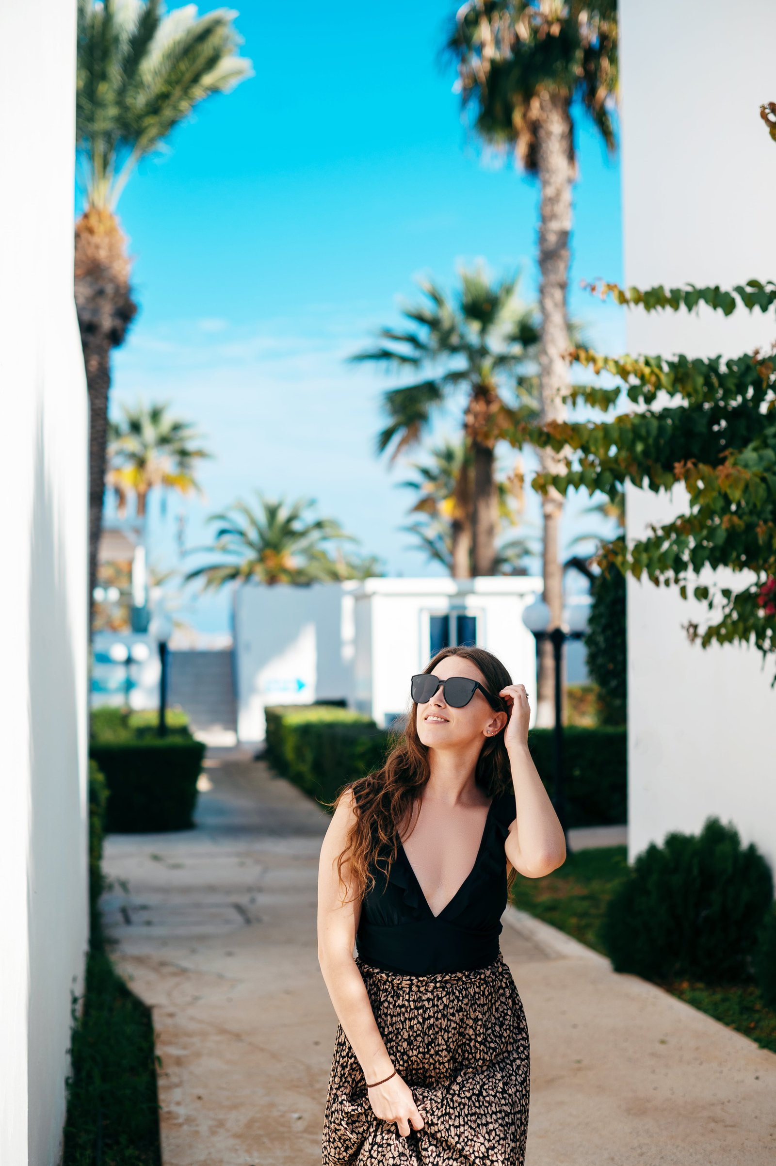femme aux cheveux noirs portant des lunettes de soleil et une robe élégante debout parmi les palmiers sur le territoire de l'hôtel complexe de luxe pour les vacances d'été
